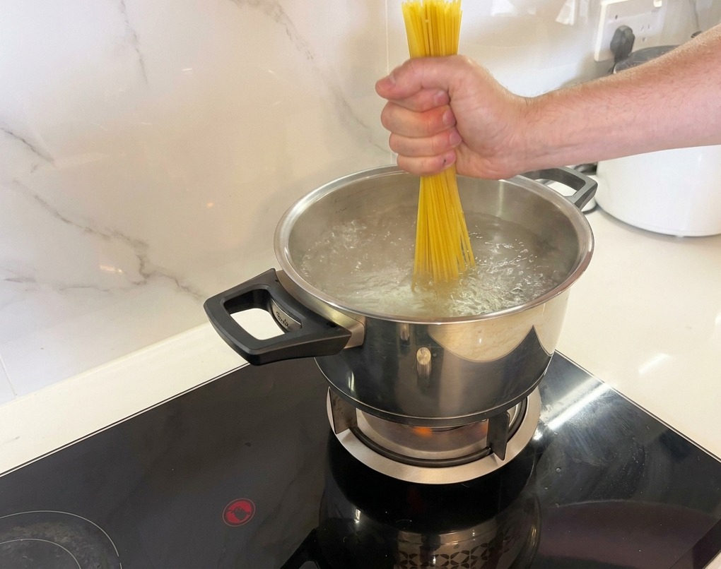 A hand pushing a bundle of dry spaghetti noodles down into a pot of vigorously boiling water.