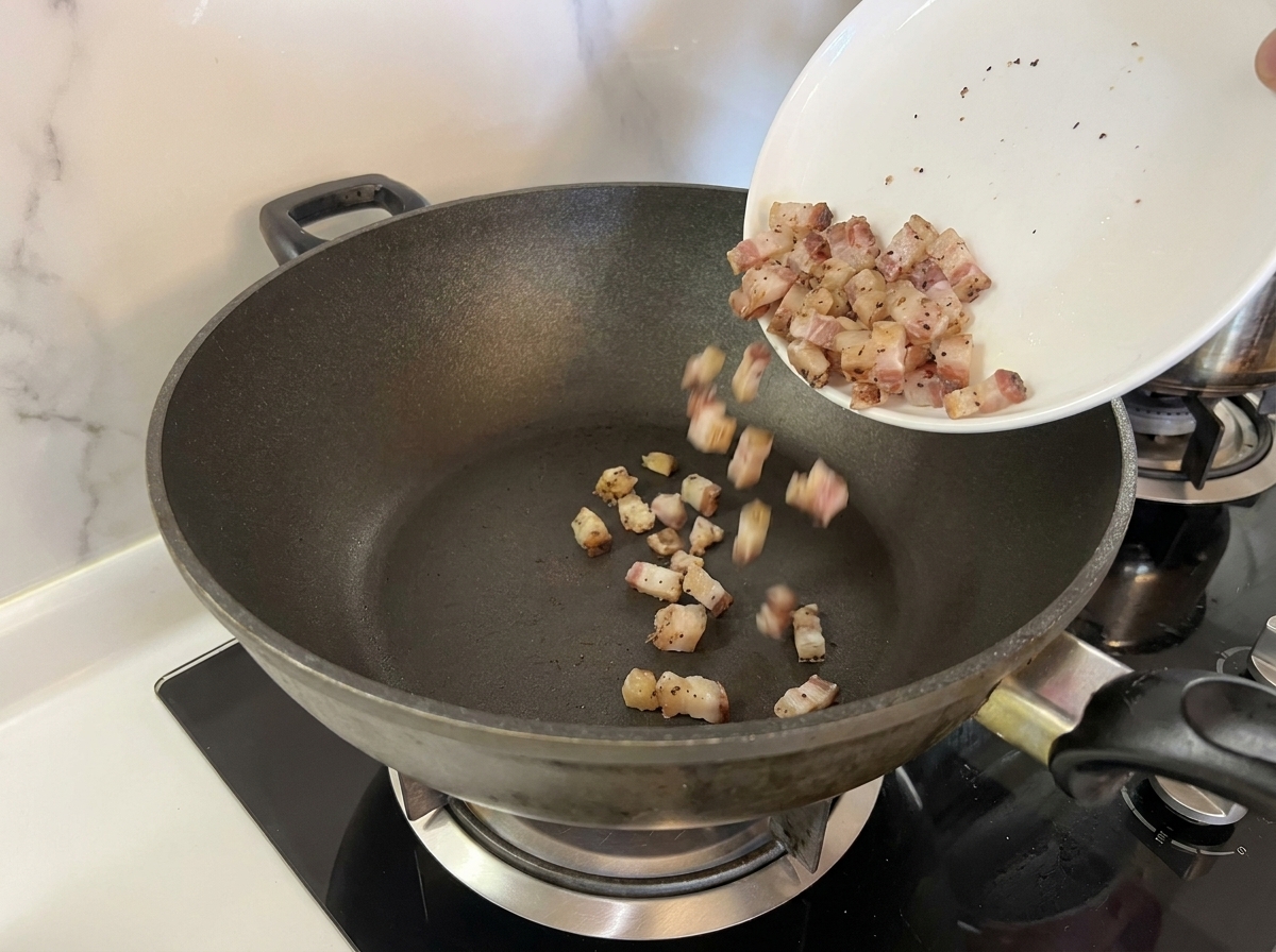 Small cubes of raw Guanciale being poured from a white plate into a dry, dark frying pan.