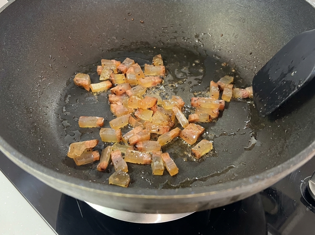 Cubes of Guanciale frying in their own rendered fat in a dark pan, turning translucent and slightly browned.