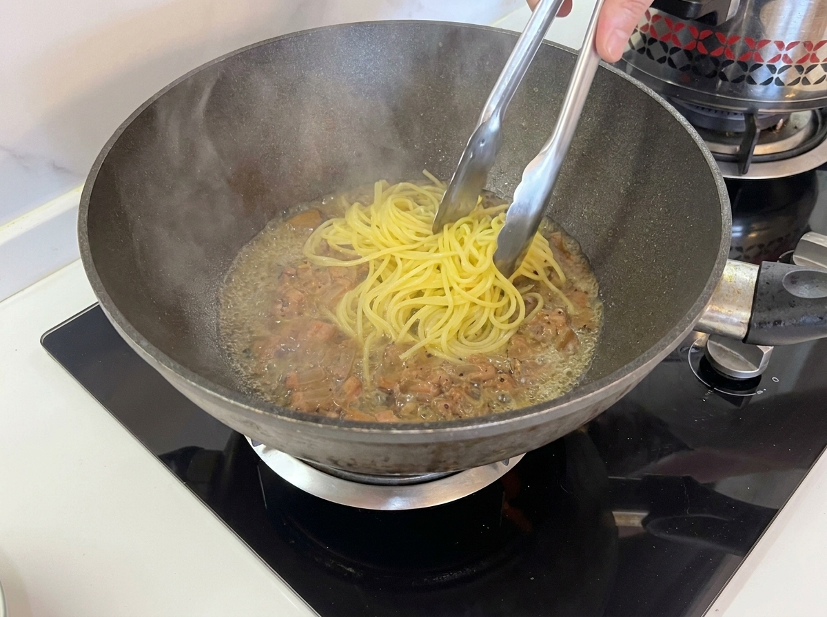 Tongs lifting cooked spaghetti from a boiling pot directly into a pan containing cooked guanciale.