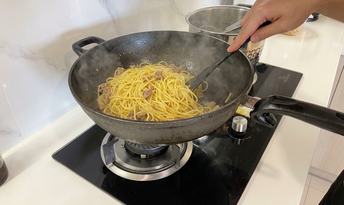 A hand vigorously stirring and tossing spaghetti in a pan to create a creamy, emulsified carbonara sauce.