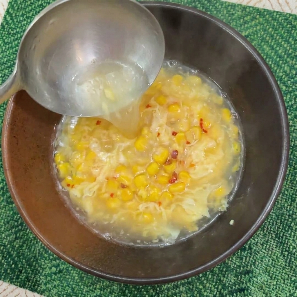 A metal ladle pouring hot, steaming water into a dark brown bowl filled with corn kernels and an egg mixture.