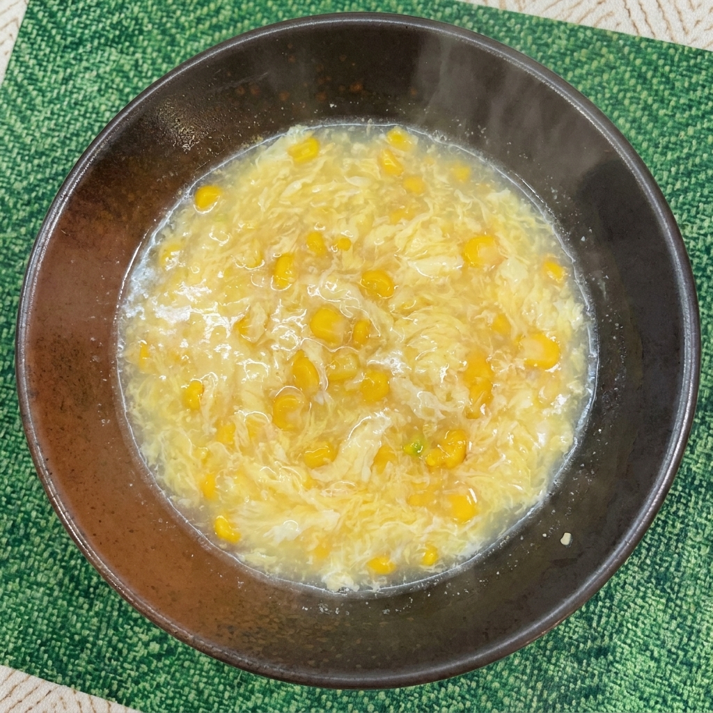 A creamy, light yellow broth mixed with sweet corn kernels and delicate egg strands in a dark serving bowl.