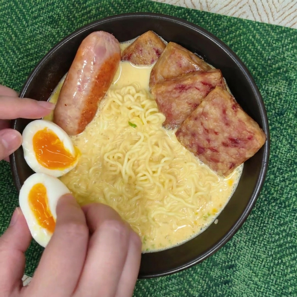 Hands carefully placing two halves of a soft-boiled egg with a bright orange yolk into a ramen bowl next to a cooked sausage and slices of spam.