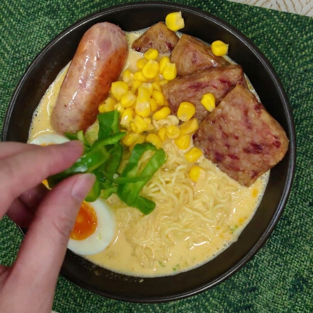 A hand placing fresh green bell pepper slices onto a bowl of creamy ramen noodles topped with corn, an egg half, and savory meats.