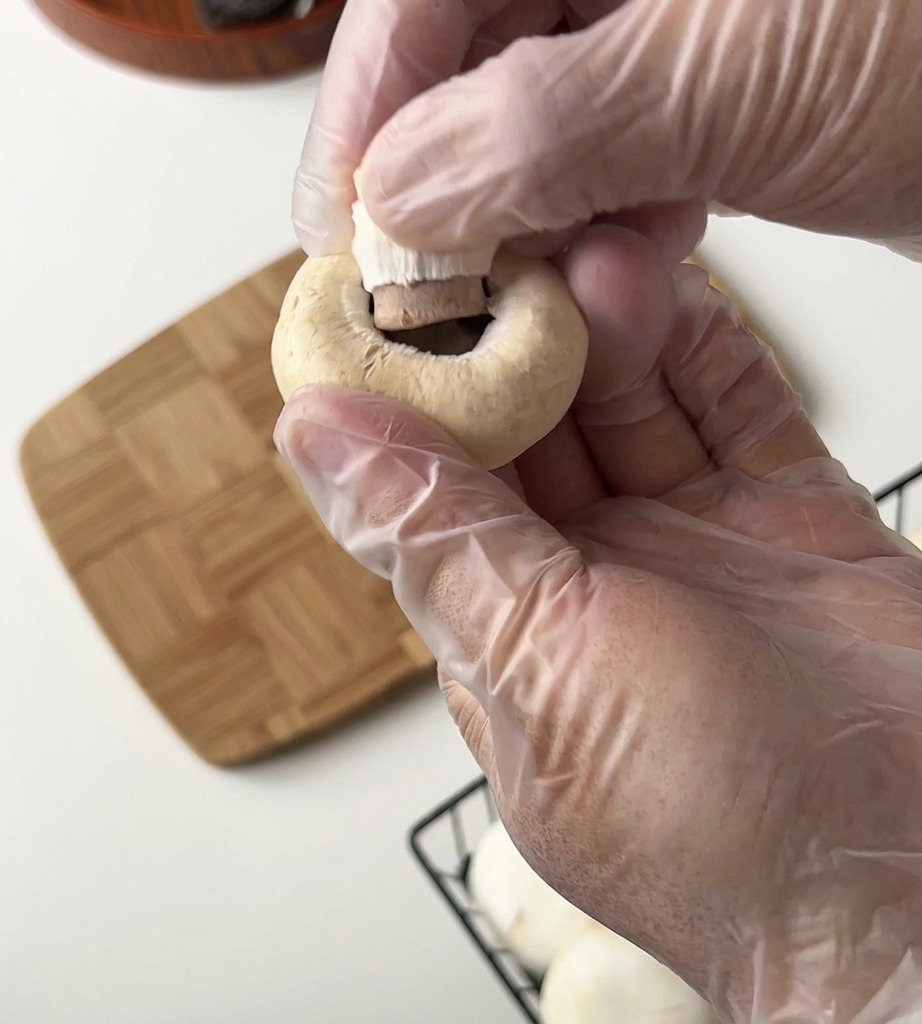 A person wearing clear plastic gloves holds a white button mushroom and pulls the stem out of the cap over a wooden board.