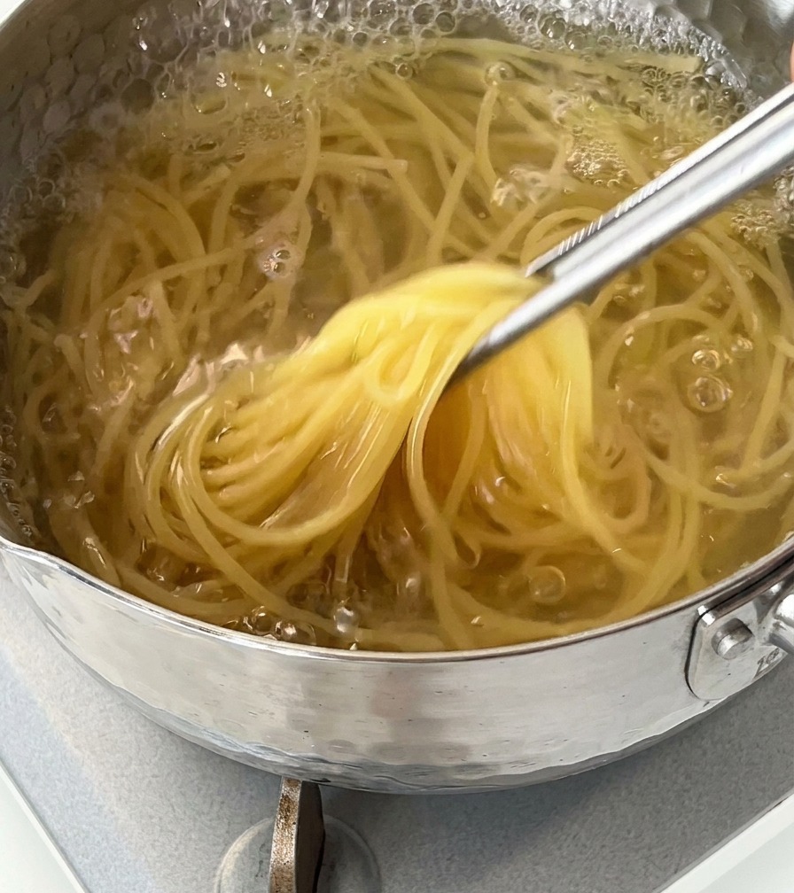 A close-up shot of spaghetti noodles being lifted out of a pot of bubbling boiling water using metal tongs.