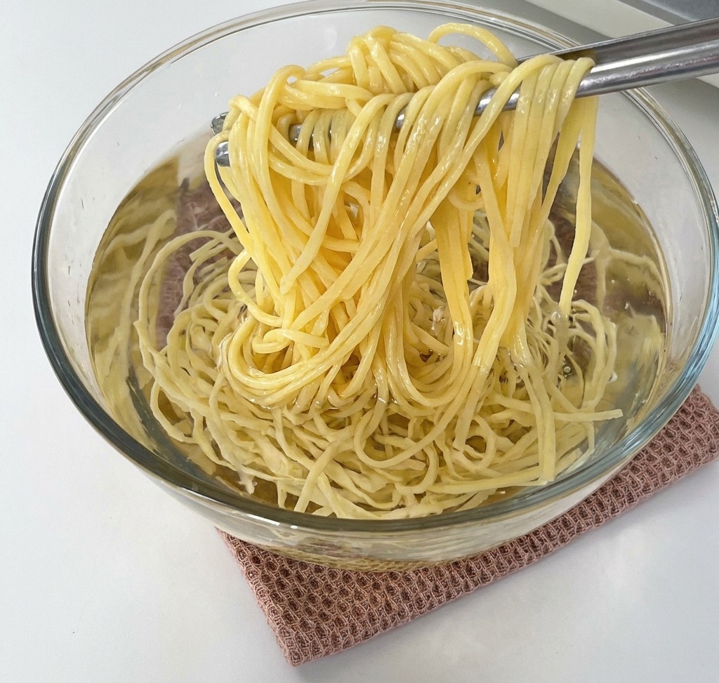 A pair of tongs lifting a bundle of pale yellow cooked spaghetti from a clear glass bowl filled with water.