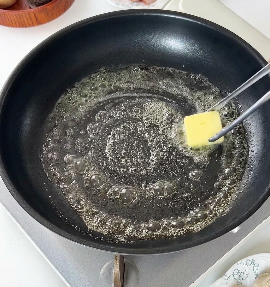 A square pat of yellow butter melting and bubbling in a black non-stick frying pan.