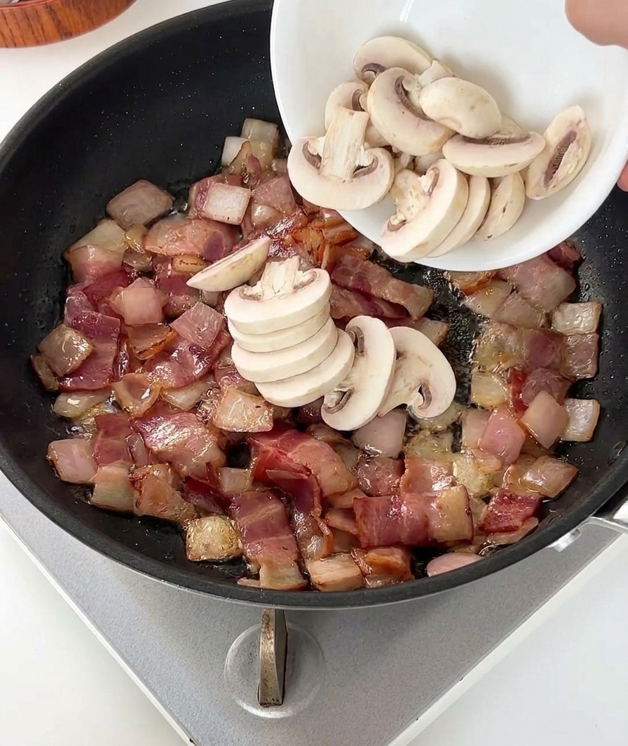 A person pouring a bowl of fresh white sliced mushrooms into a black frying pan containing browned bacon bits and translucent onions.