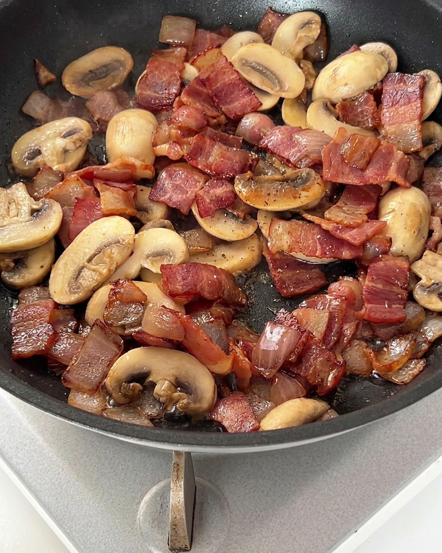 A pan containing browned bacon, sautéed onions, and sliced mushrooms sprinkled with black pepper.
