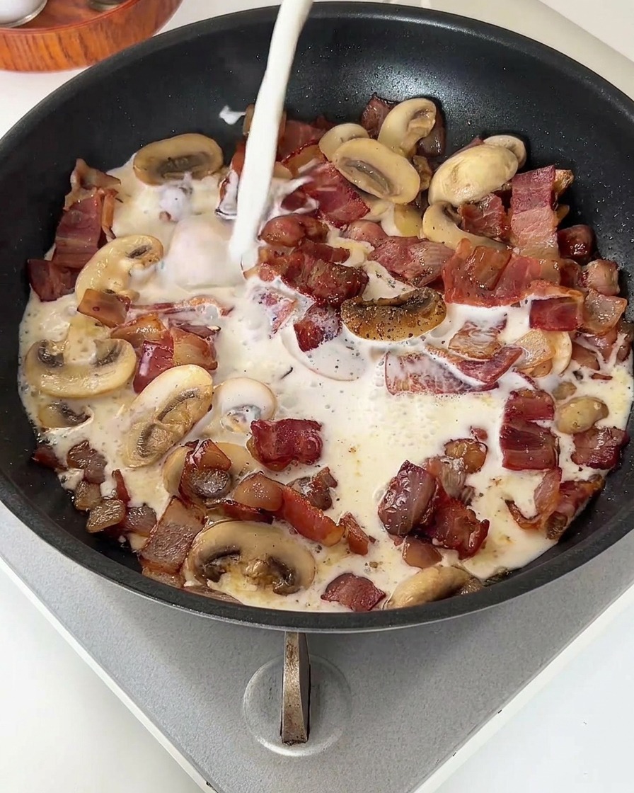A stream of white milk being poured into a pan filled with cooked bacon and mushrooms.