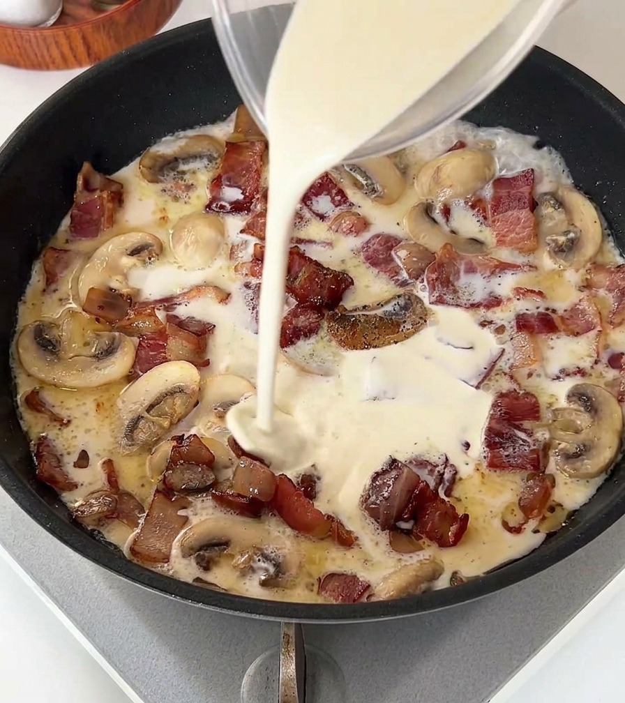 A stream of white light cream being poured from a glass measuring cup into a pan containing cooked bacon and sliced mushrooms.