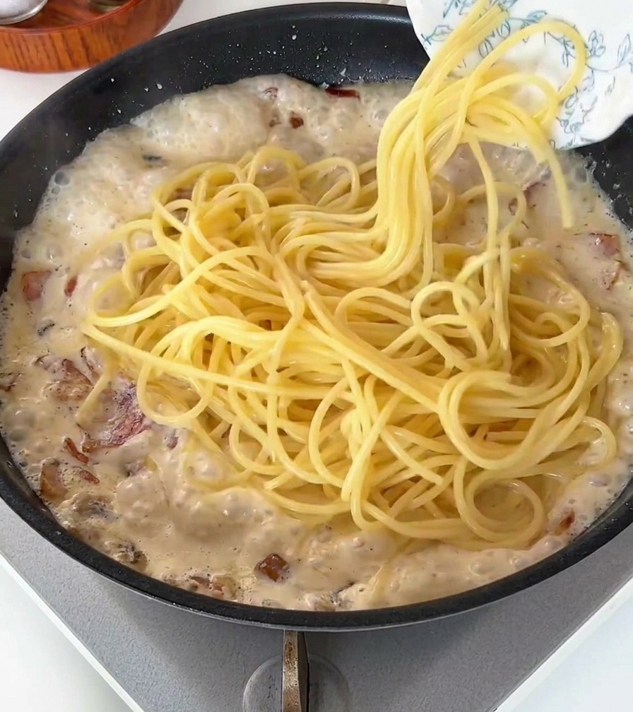 Cooked spaghetti noodles being dropped into a pan of white, bubbling creamy sauce with bacon.