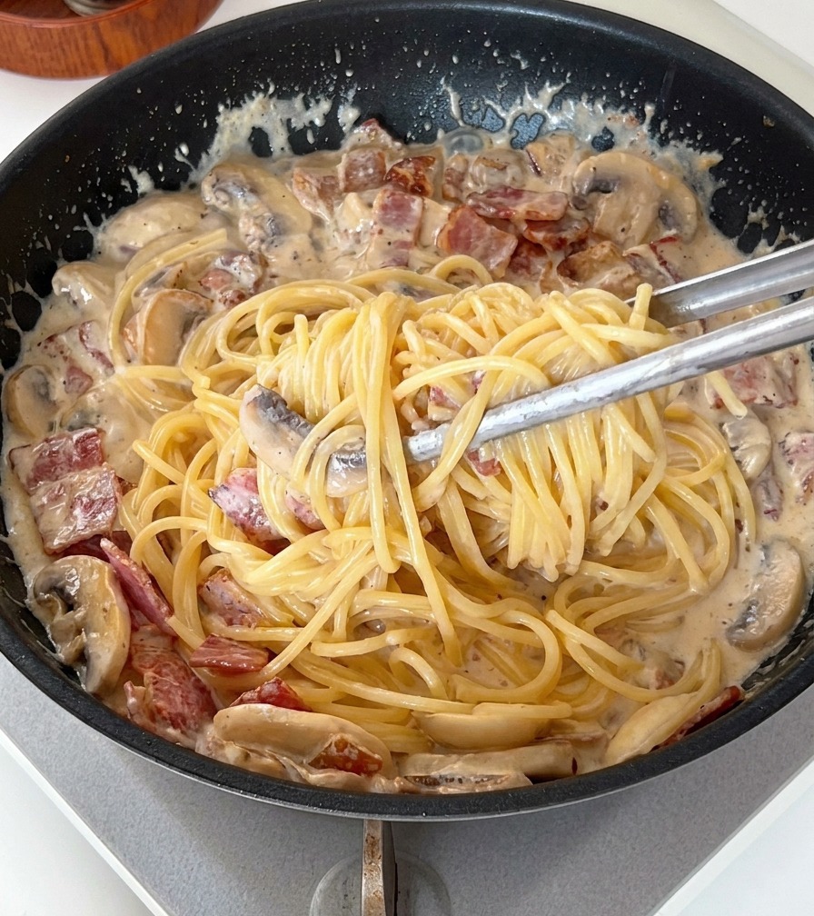 Tongs lifting and mixing spaghetti noodles within a thick, creamy sauce filled with bacon and mushroom slices.