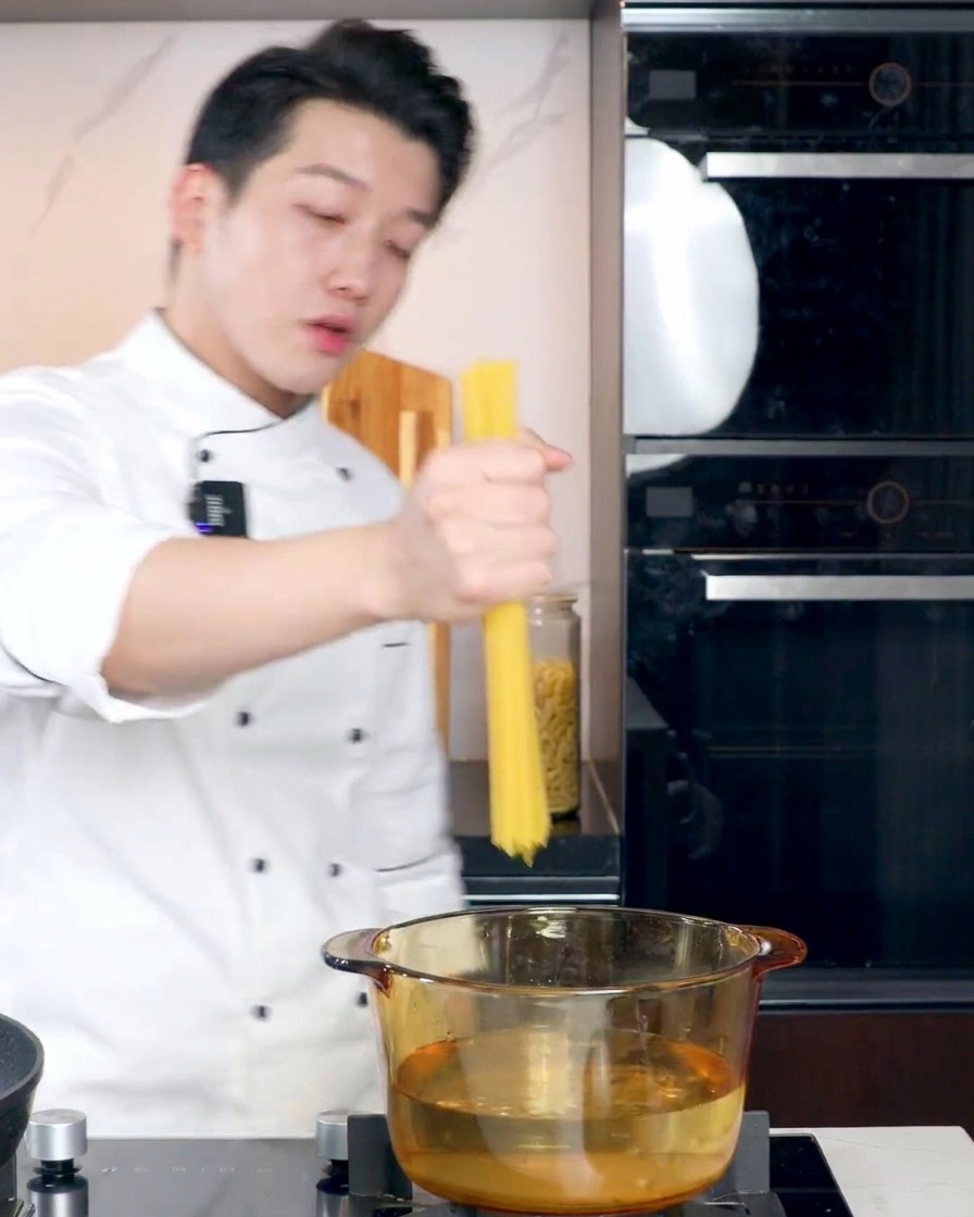 A chef in a white uniform holding a bundle of dry spaghetti over a transparent glass pot filled with boiling water on a stovetop.