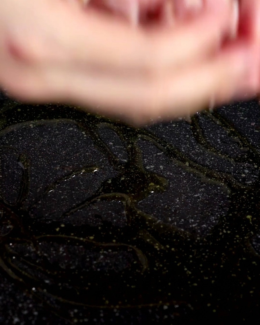 Close-up of raw bacon pieces being dropped into a dark frying pan coated with shimmering olive oil.