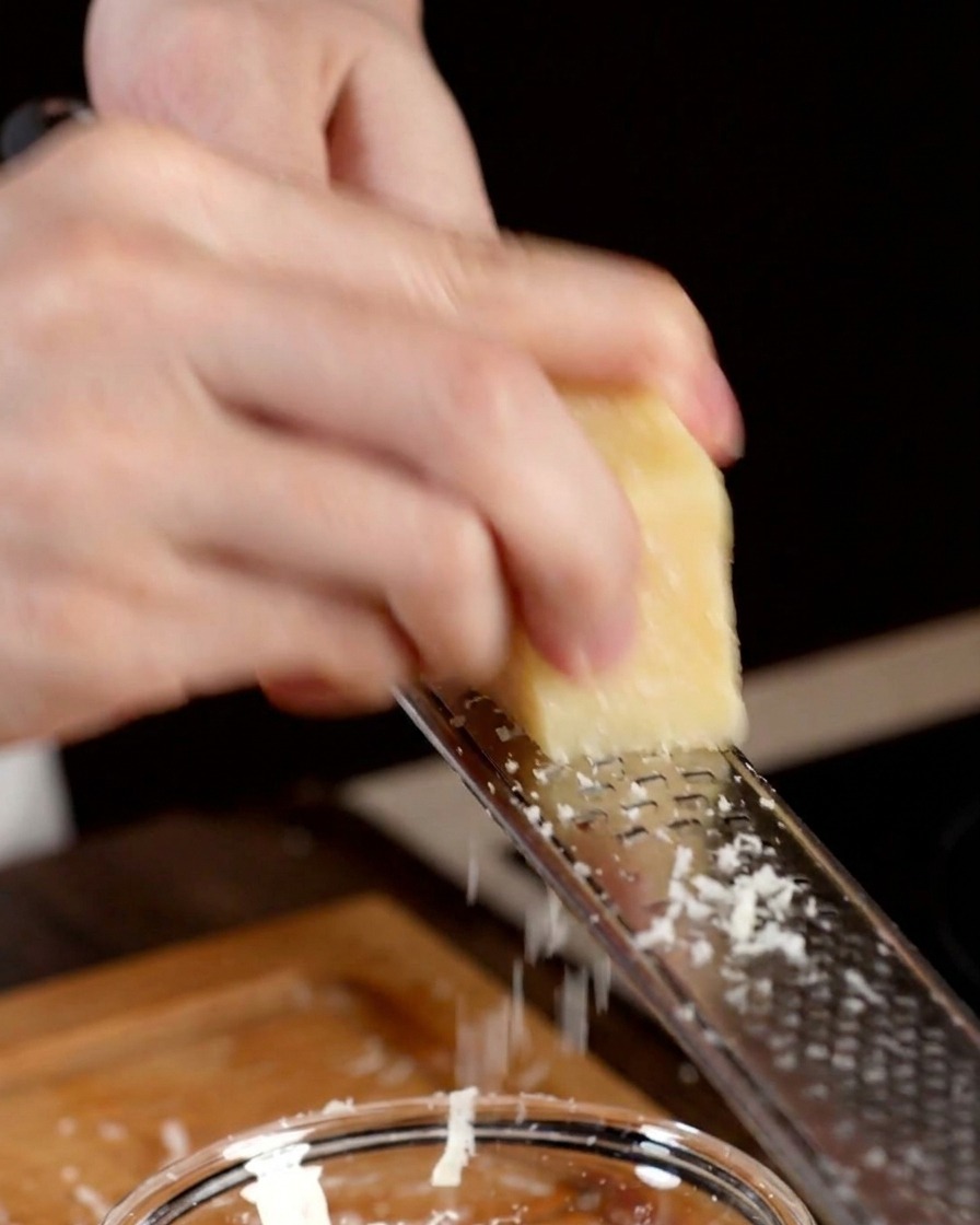 A hand using a metal grater to shred a wedge of hard Parmesan cheese into a bowl.