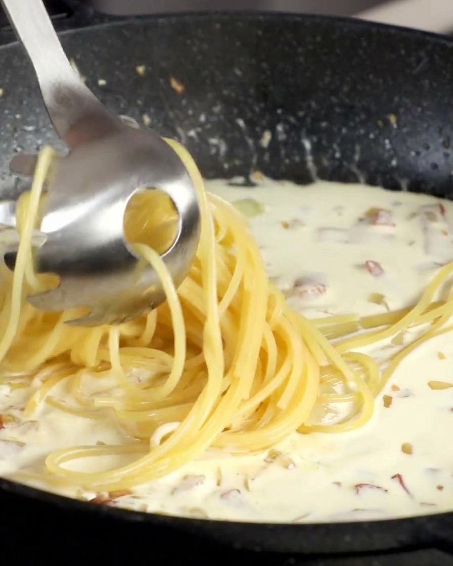 A metal pasta fork lifting a bundle of cooked spaghetti and dropping it into a bubbling white cream sauce in a black pan.