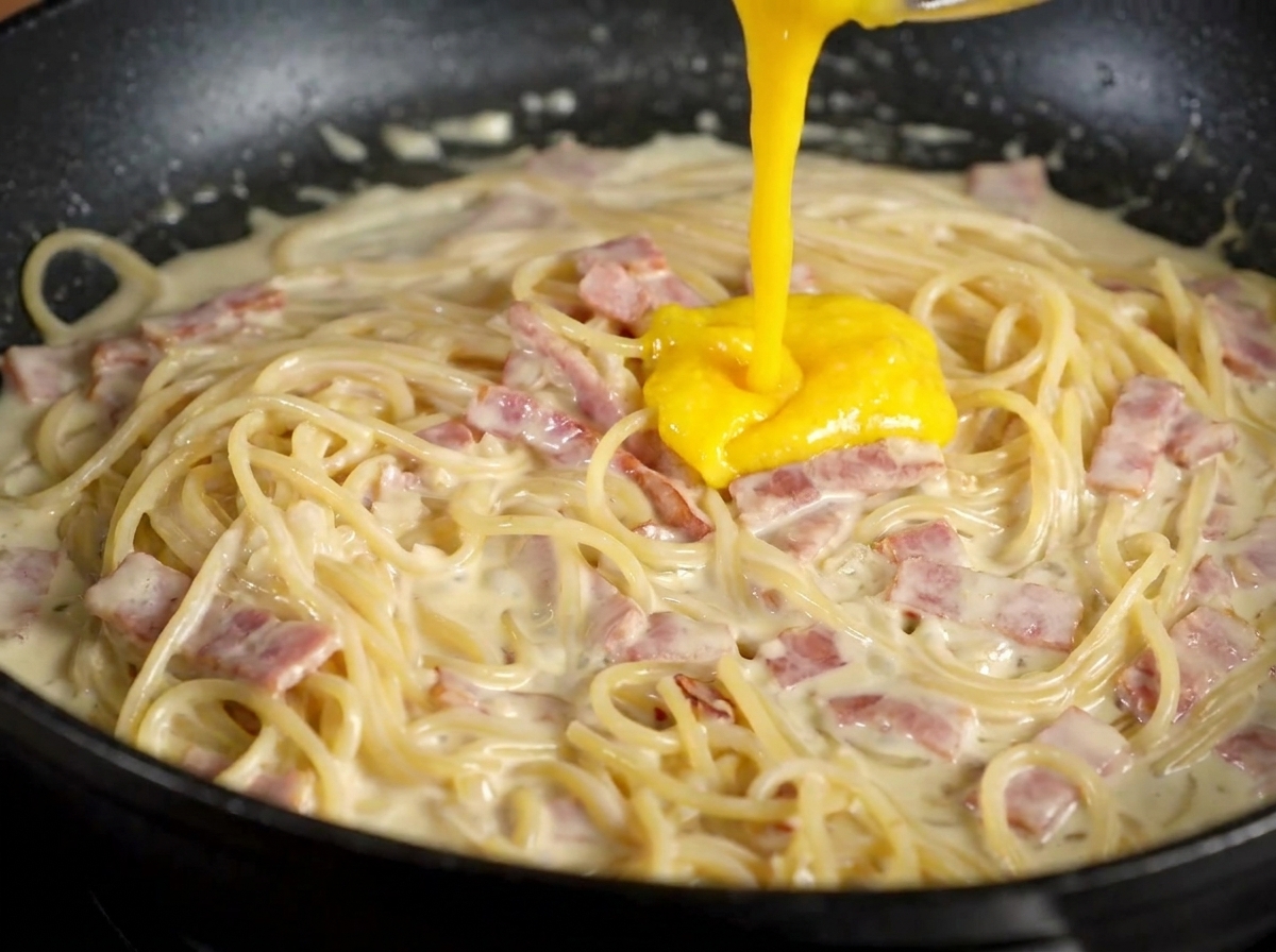 A thick, vibrant yellow egg yolk and cheese mixture being poured over creamy pasta and bacon in a black skillet.