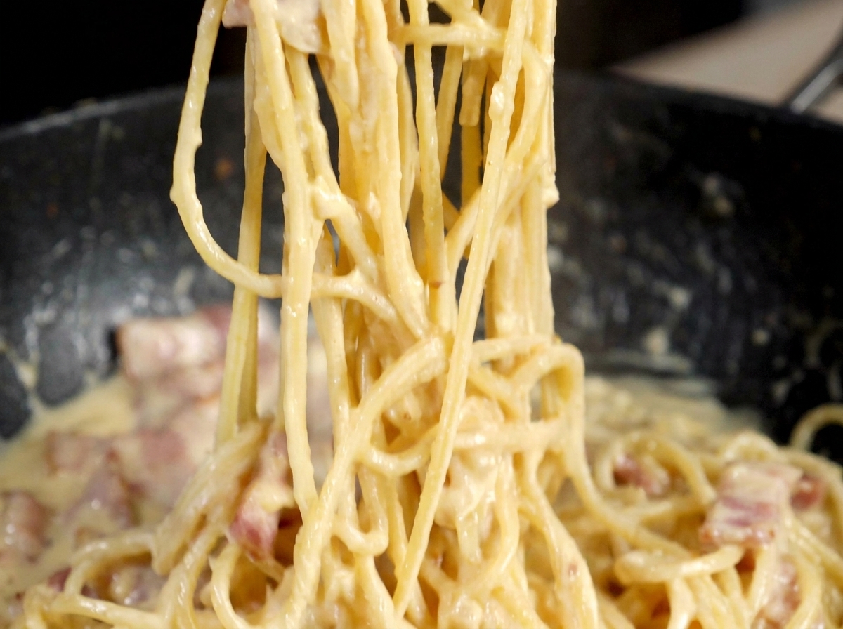 A metal pepper grinder dispensing black pepper over a pan of creamy carbonara style pasta.