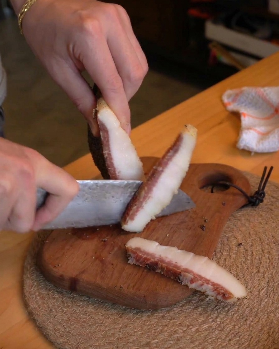 A person using a chefs knife to slice thick pieces of cured guanciale into small batons on a rustic wooden cutting board.