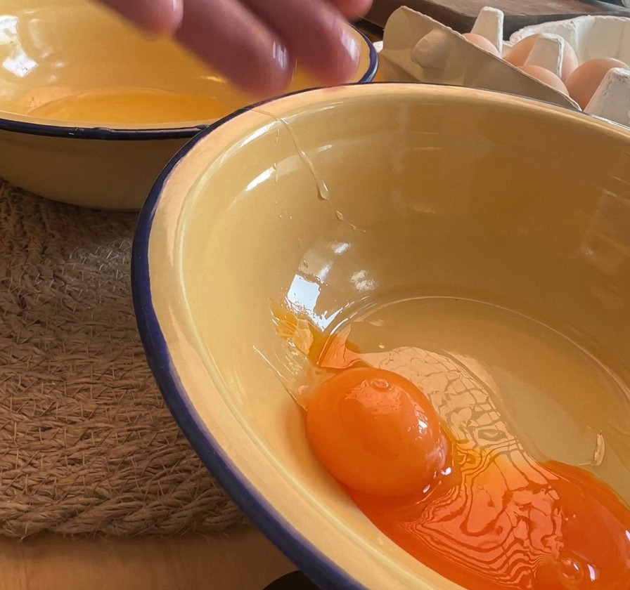 A close-up shot of a bright orange egg yolk being dropped into a yellow enamel bowl, with egg shells visible in the background.