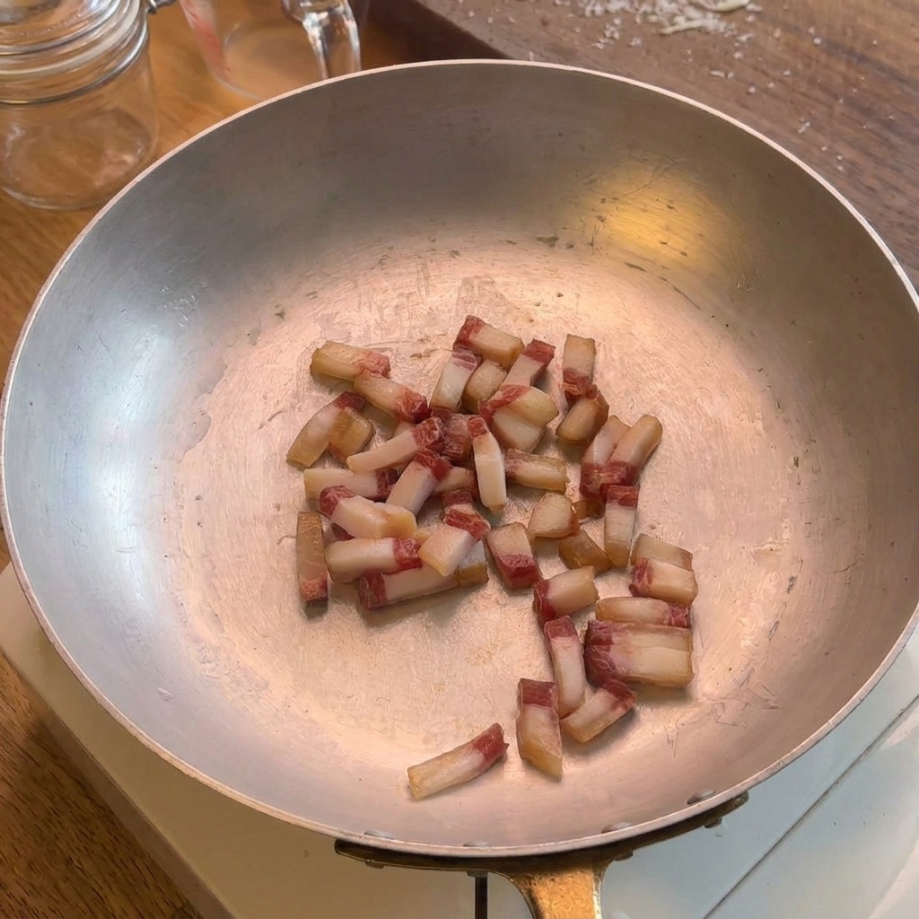 Small cubes of guanciale frying in a wide stainless steel pan, showing the natural fat beginning to render out.