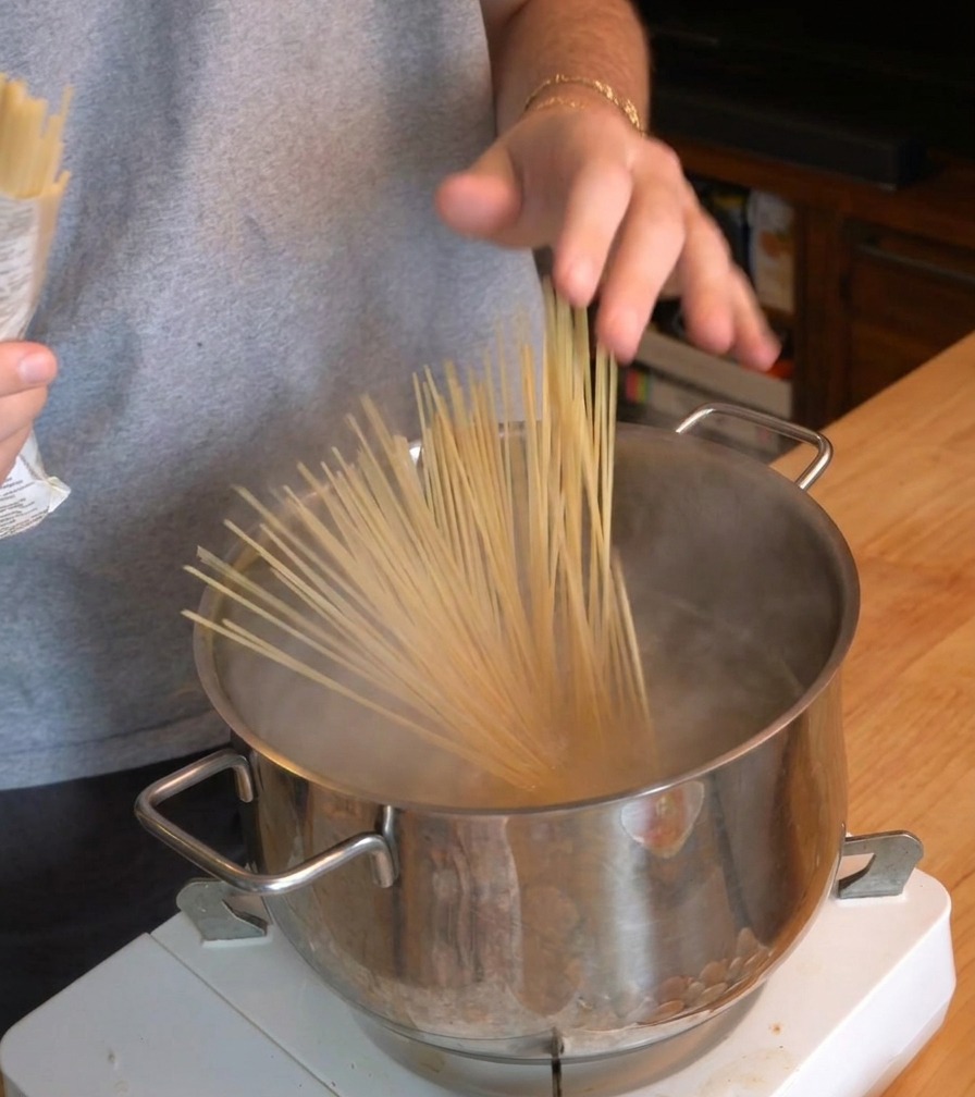 A person dropping a bundle of dry spaghetti into a steaming pot of boiling water on a portable stove.