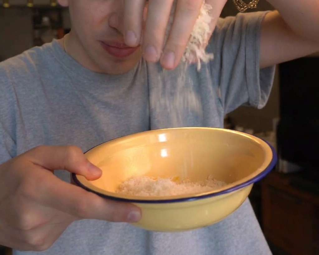 A man adding a handful of finely grated white cheese into a yellow bowl containing egg yolks.