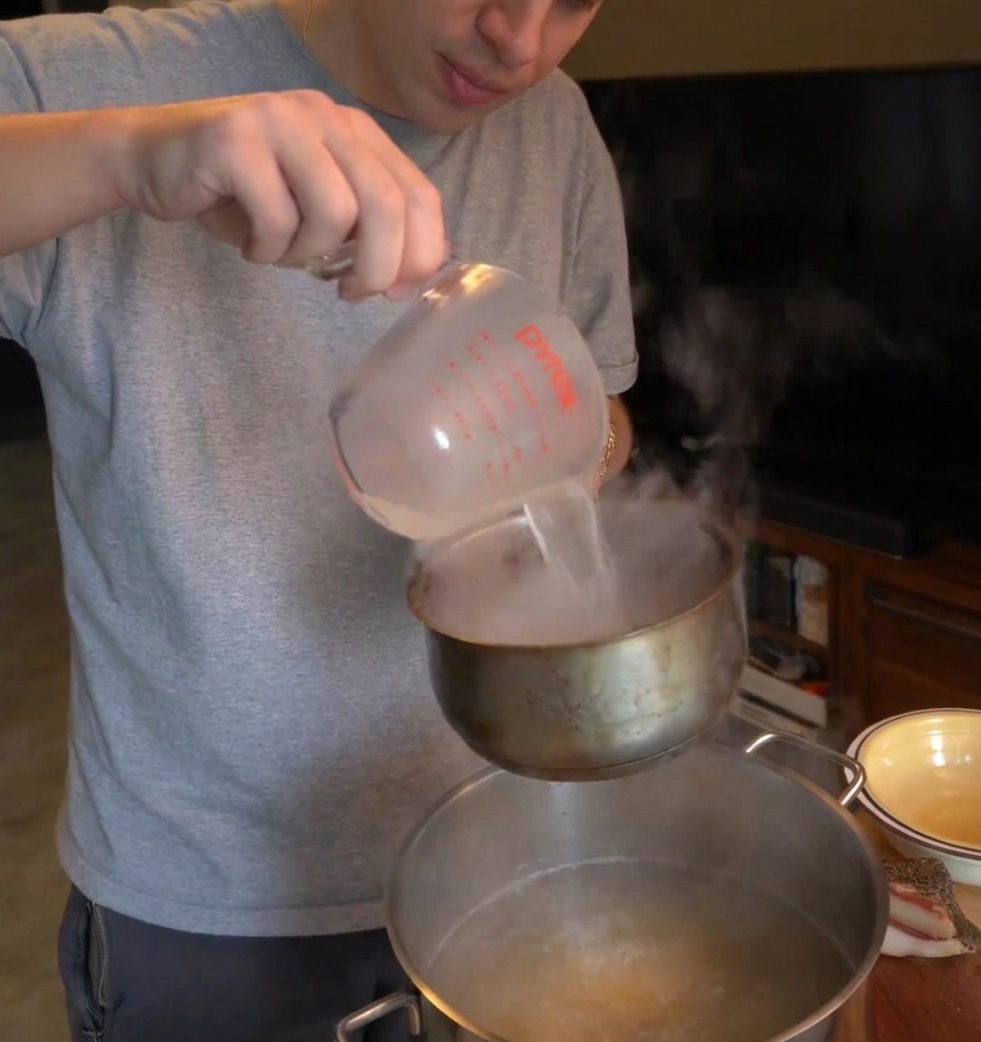 A person pouring hot, cloudy pasta water from a measuring cup into a smaller pot held over the boiling pasta water.