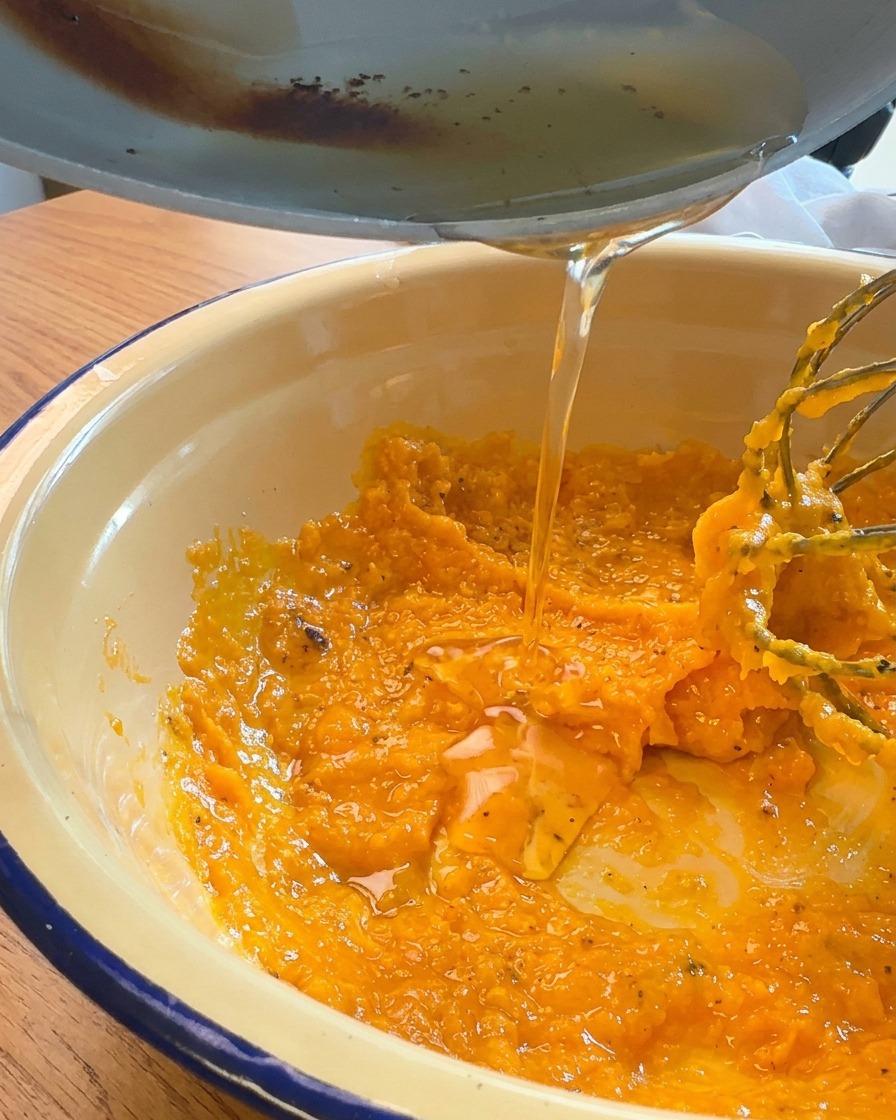 Golden liquid pork fat being poured from a pan into a bowl of thick orange egg yolk and grated cheese mixture.