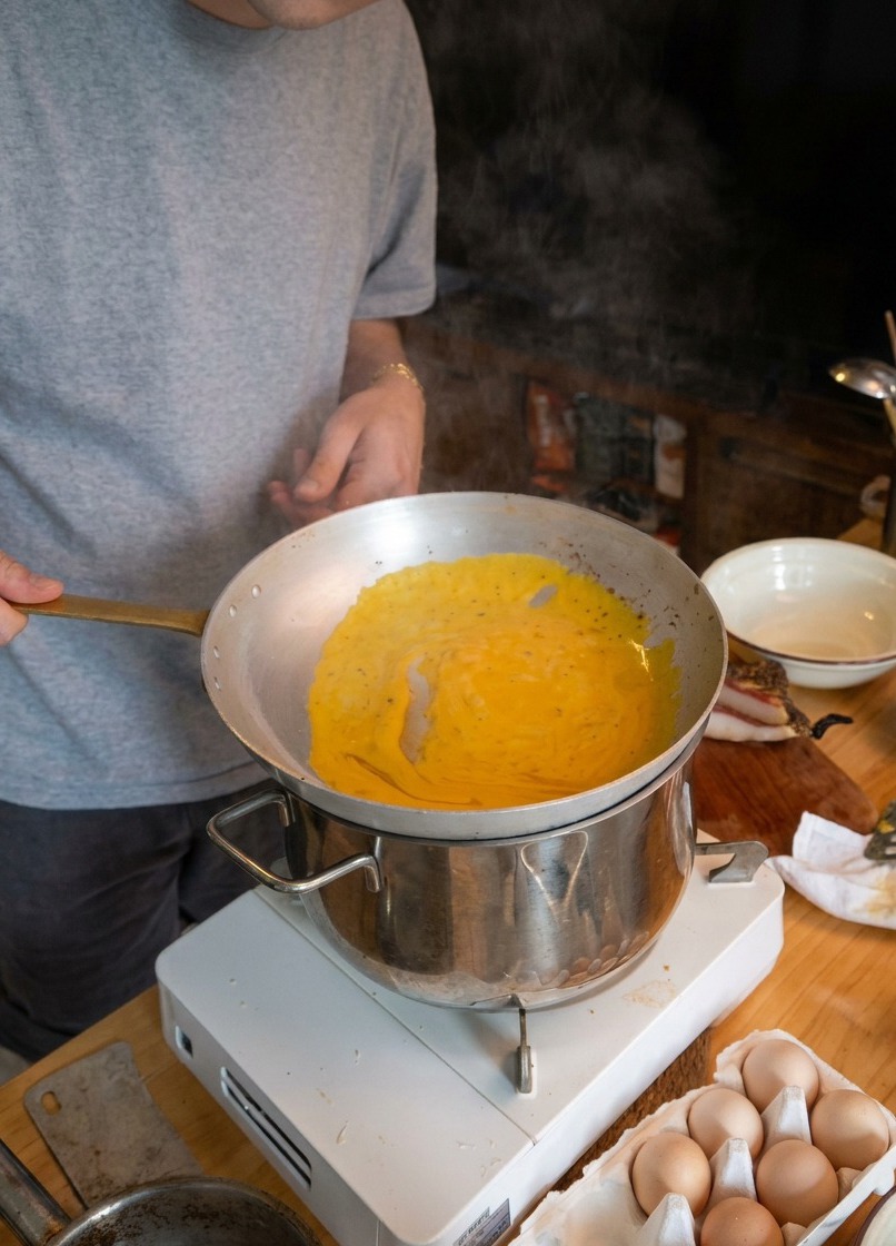 A chef tossing spaghetti in a light metal pan held over a steaming pot of water to emulsify the sauce.