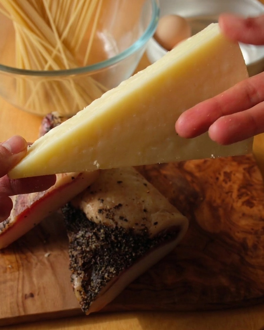 A close-up shot of a hand holding a wedge of pale Pecorino Romano cheese over two thick slabs of black-pepper-crusted guanciale on a wooden surface.