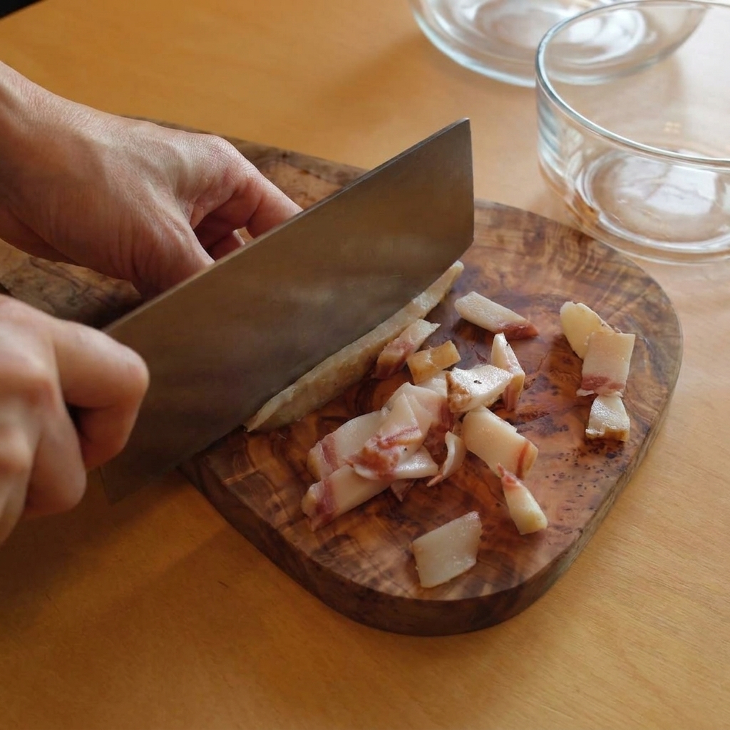 A person using a large kitchen knife to cut strips of fatty guanciale on a rustic wooden cutting board.