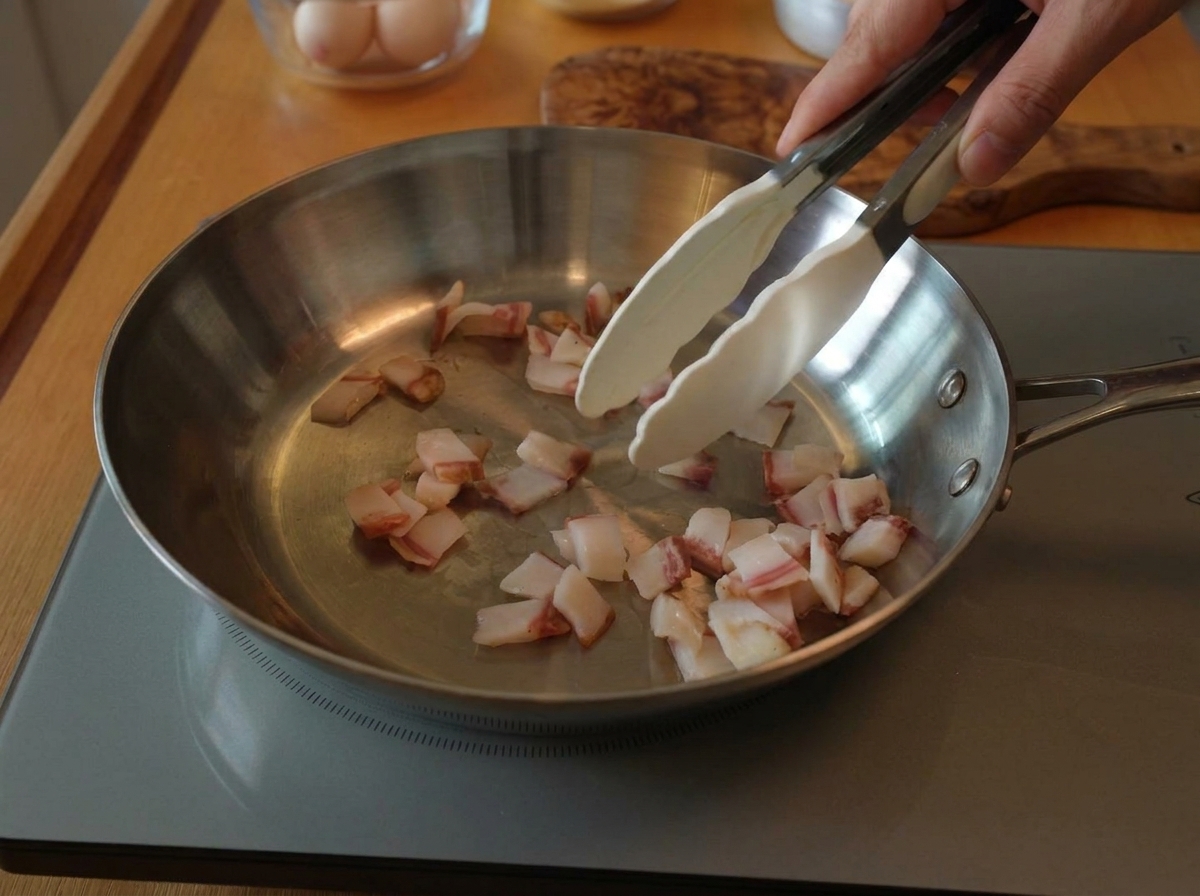 Tongs stirring small pieces of raw guanciale in a wide stainless steel skillet over an induction cooktop.