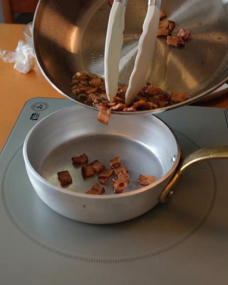 Crispy browned guanciale pieces being poured from a stainless steel pan into a small silver bowl using white-tipped tongs.