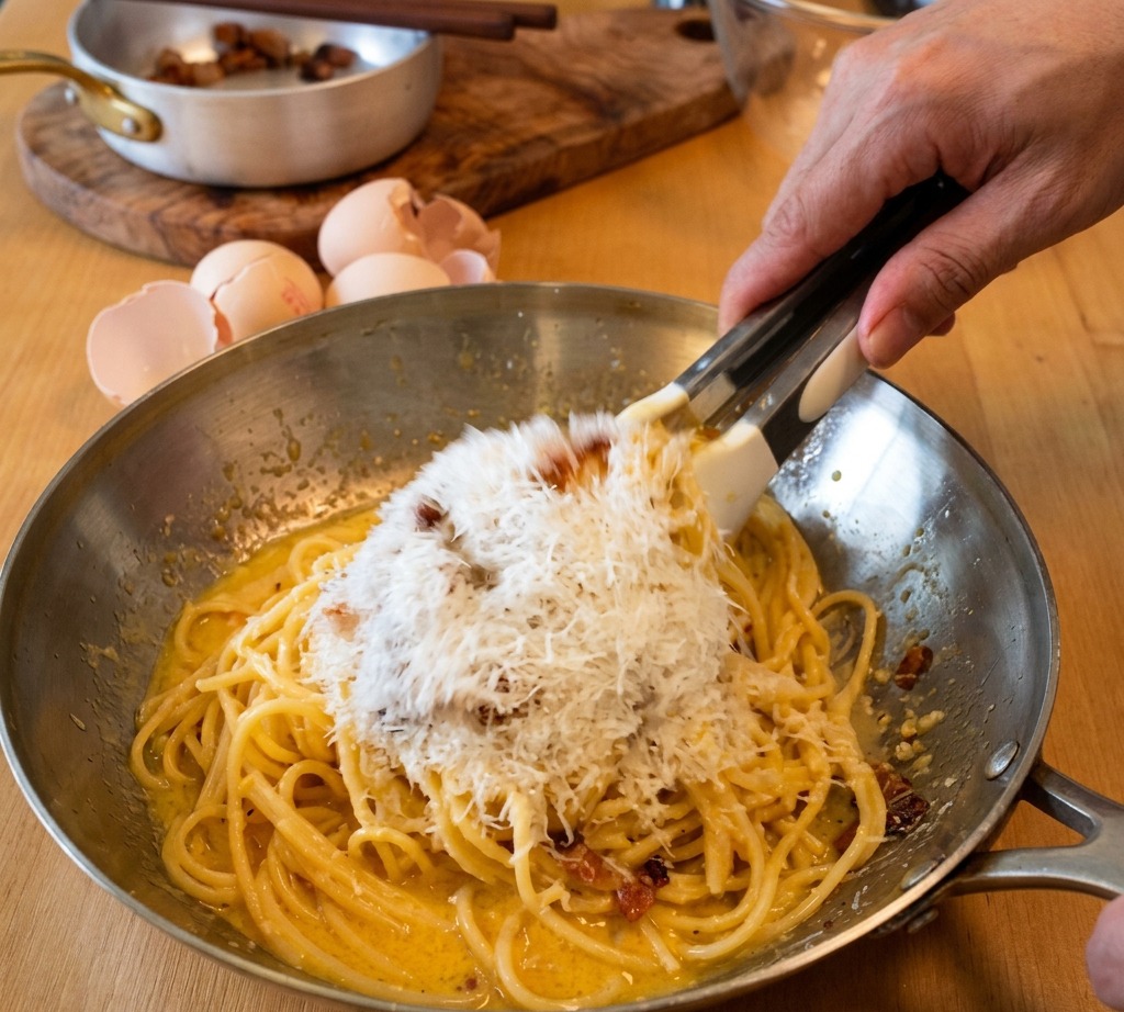 Tongs tossing spaghetti in a metal pan while a large mound of finely grated Pecorino Romano cheese is added to the creamy egg sauce.