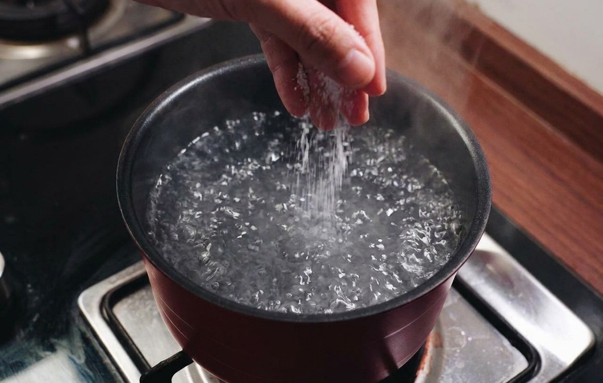 A hand sprinkling salt into a pot of vigorously boiling water on a kitchen stove.