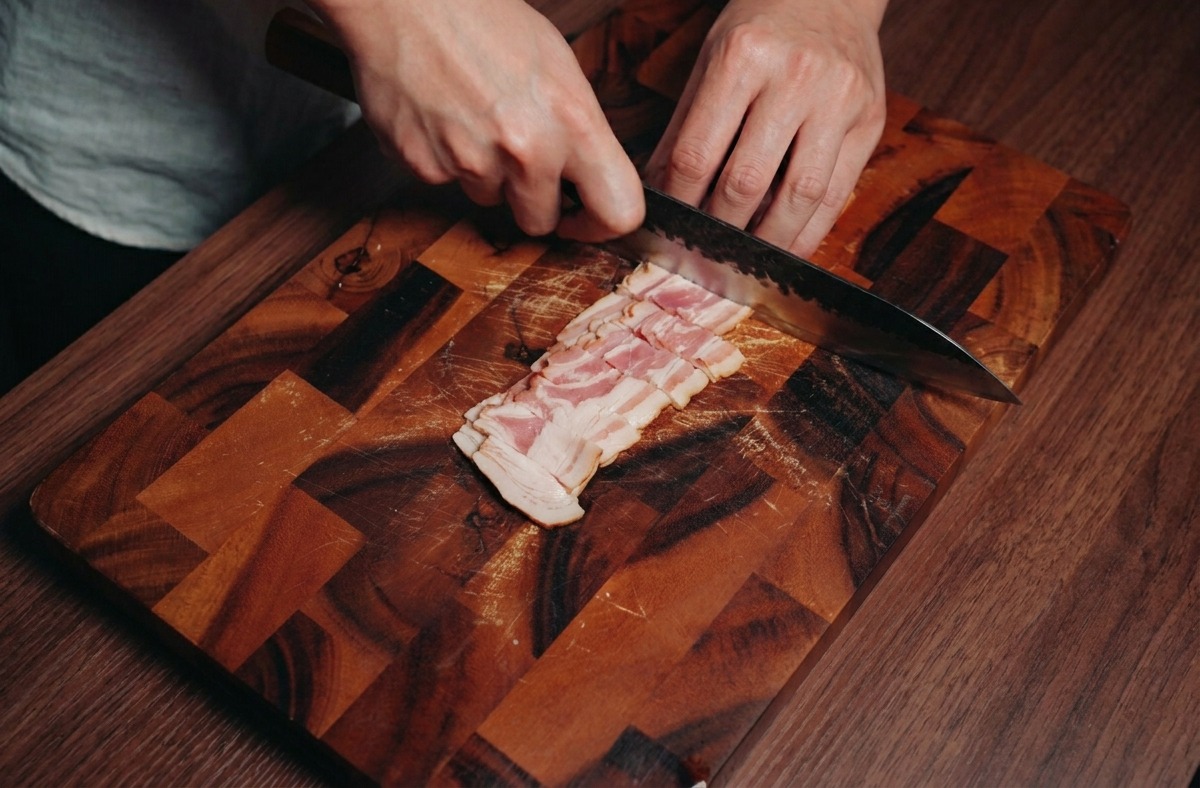 A person using a chefs knife to slice several strips of bacon into small squares on a dark wood cutting board.