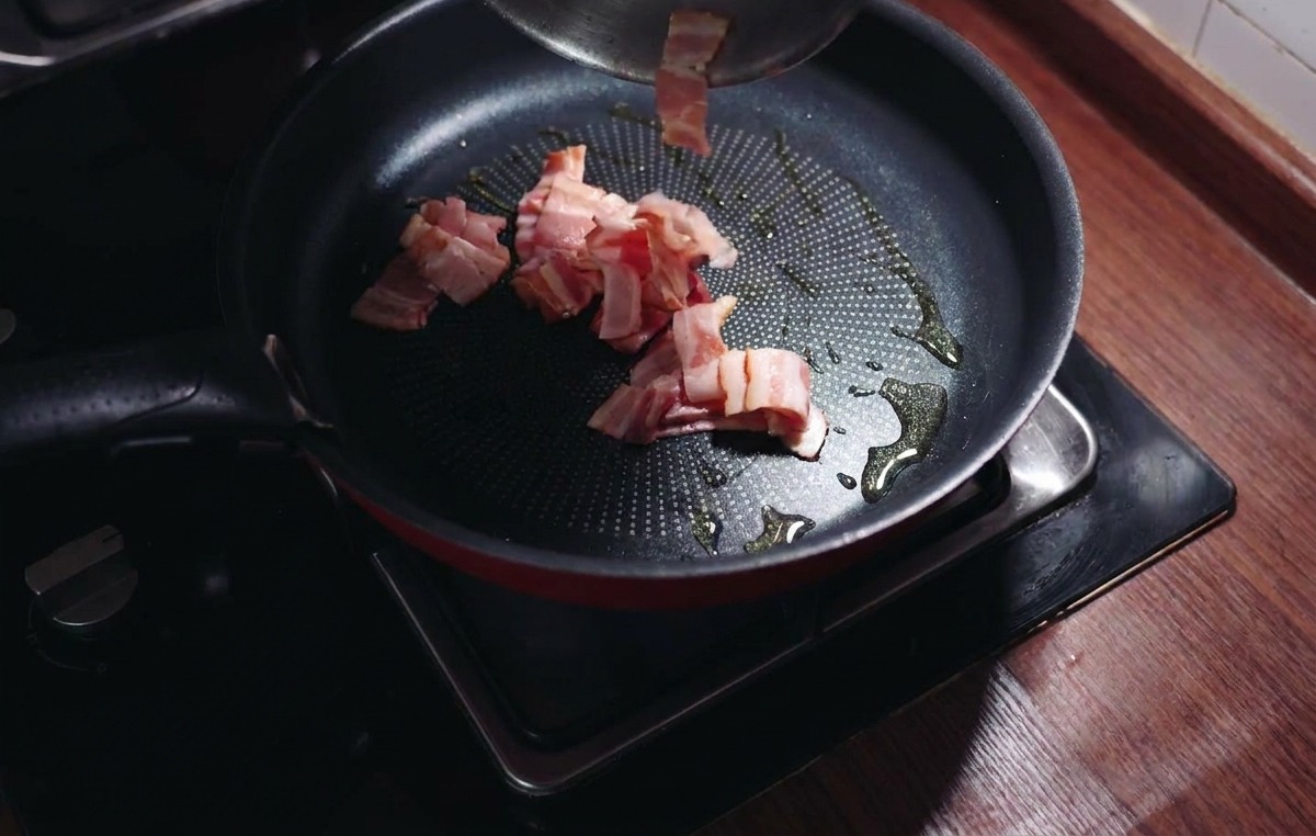 Chopped bacon pieces being added to a hot frying pan with shimmering olive oil.