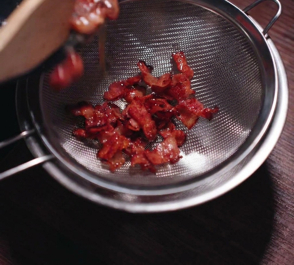 Crispy fried bacon being poured into a stainless steel mesh strainer to separate the liquid fat.