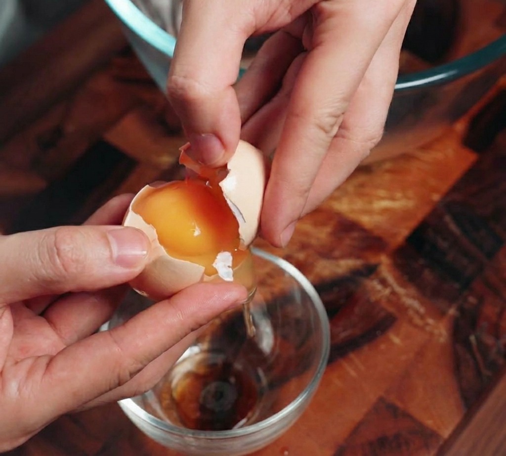 A close-up of hands cracking an egg and separating the bright orange yolk from the clear egg white.