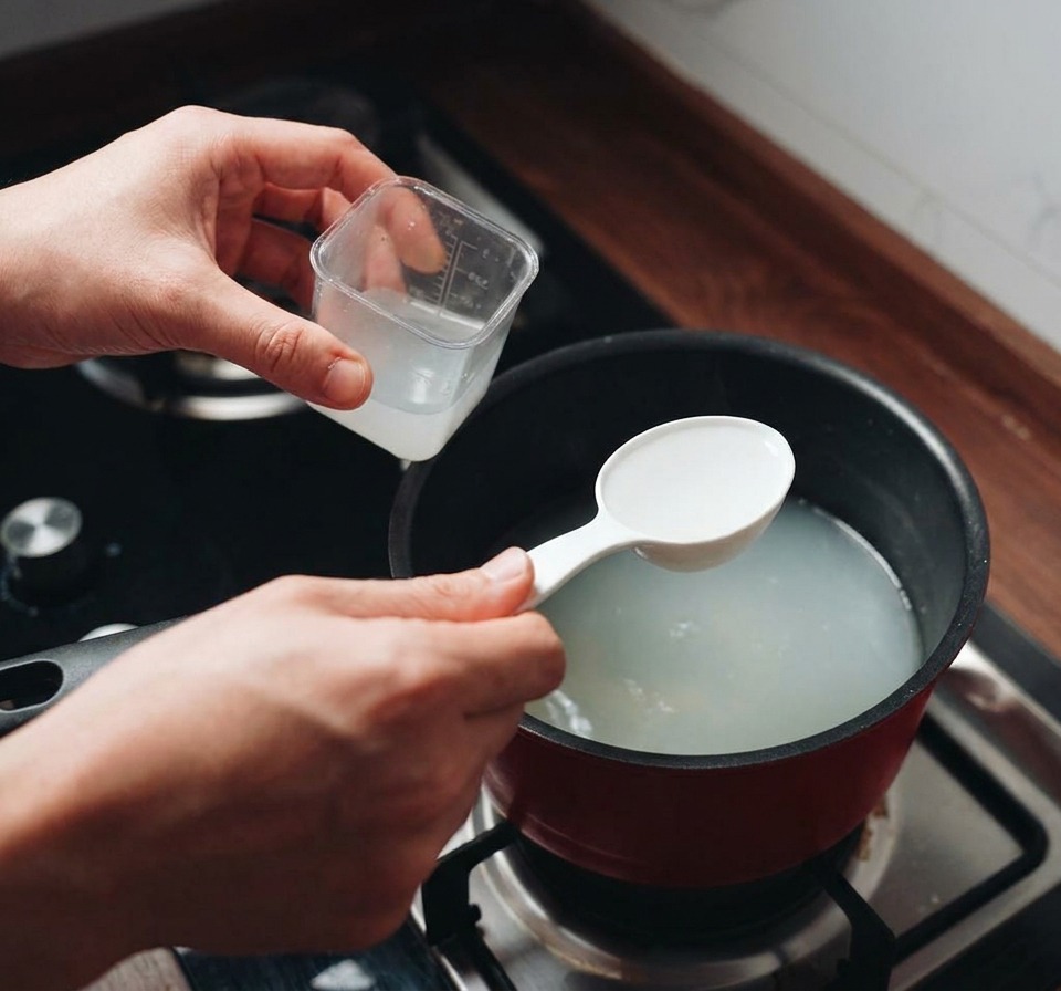 A person using a white measuring scoop to take cloudy pasta water from a simmering red pot on the stove.