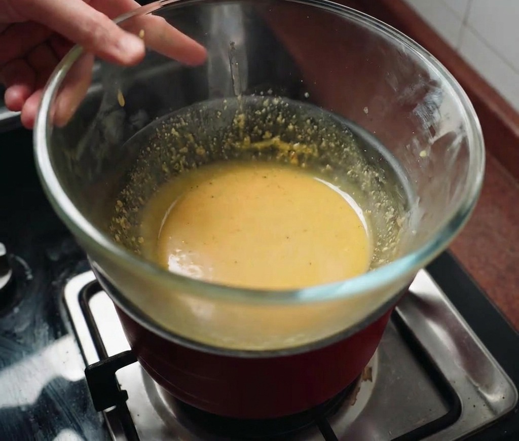 A glass bowl of yellow sauce being held over a steaming red pot of water on a gas range.