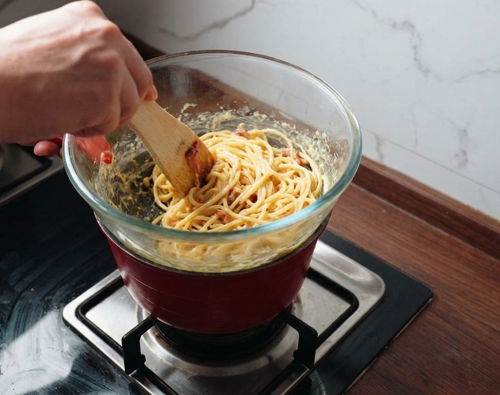 A person using a spatula to mix pasta and bacon into a creamy golden sauce inside a glass bowl held over a steaming pot