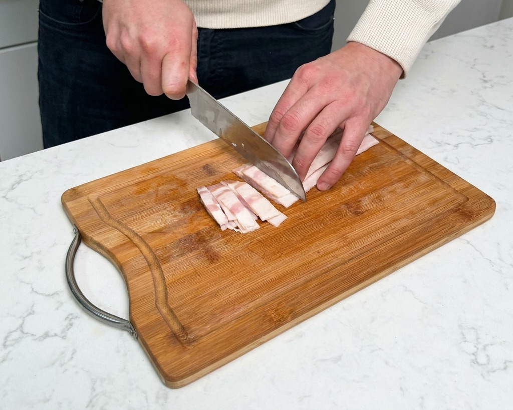 Hand slicing raw bacon strips into small rectangles on a wooden cutting board.