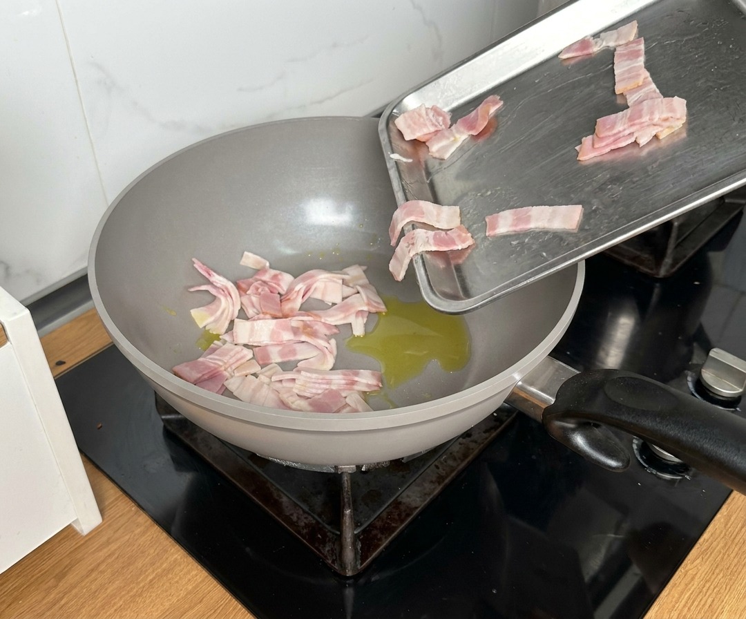 Bacon pieces being dropped from a metal tray into a large pan with shimmering oil.