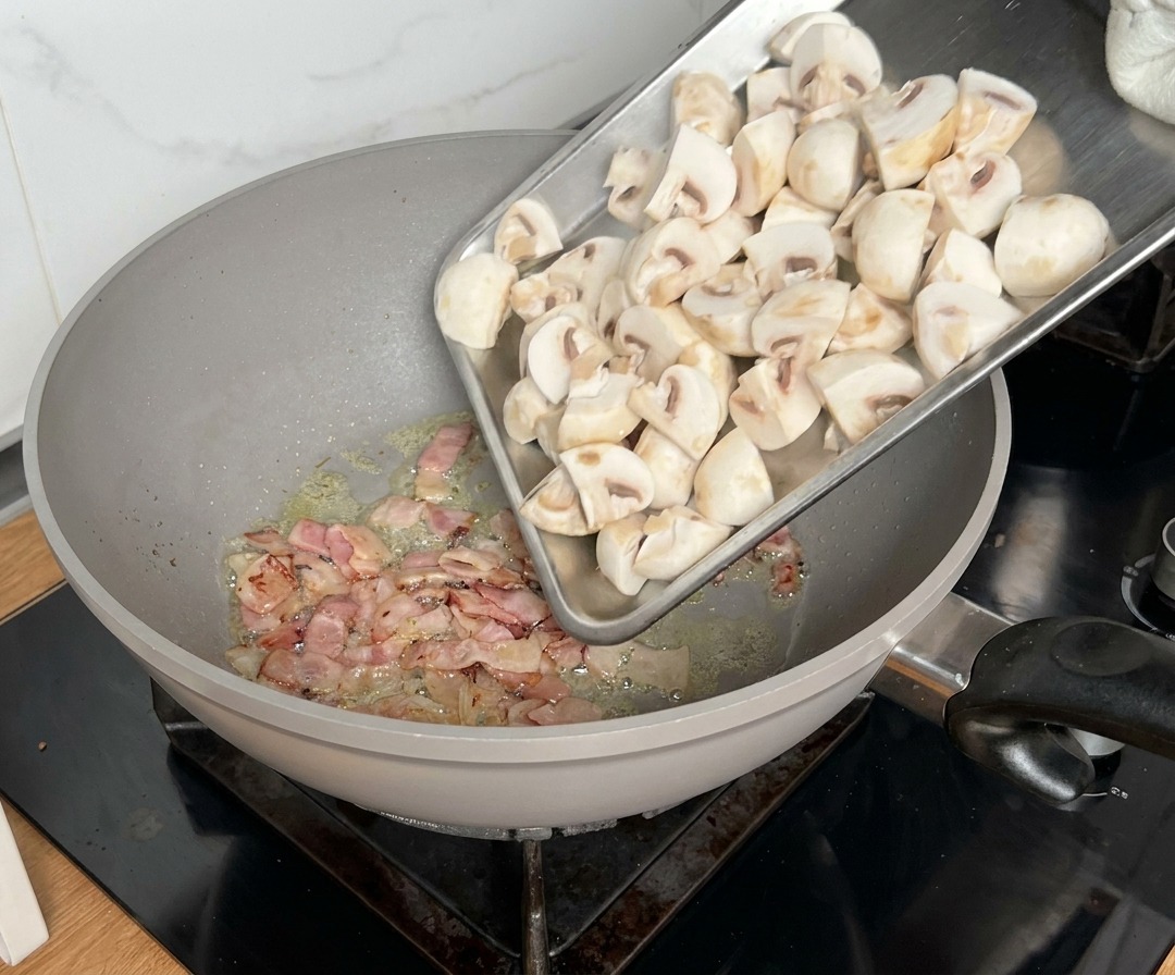 Quartered mushrooms being added to a pan where bacon is already cooking.