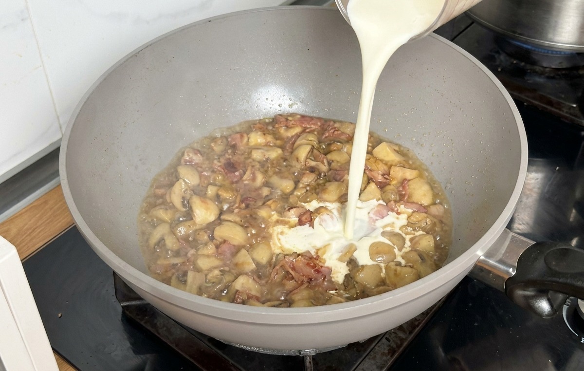 Heavy cream being poured from a measuring cup into a pan with simmering stock and mushrooms.